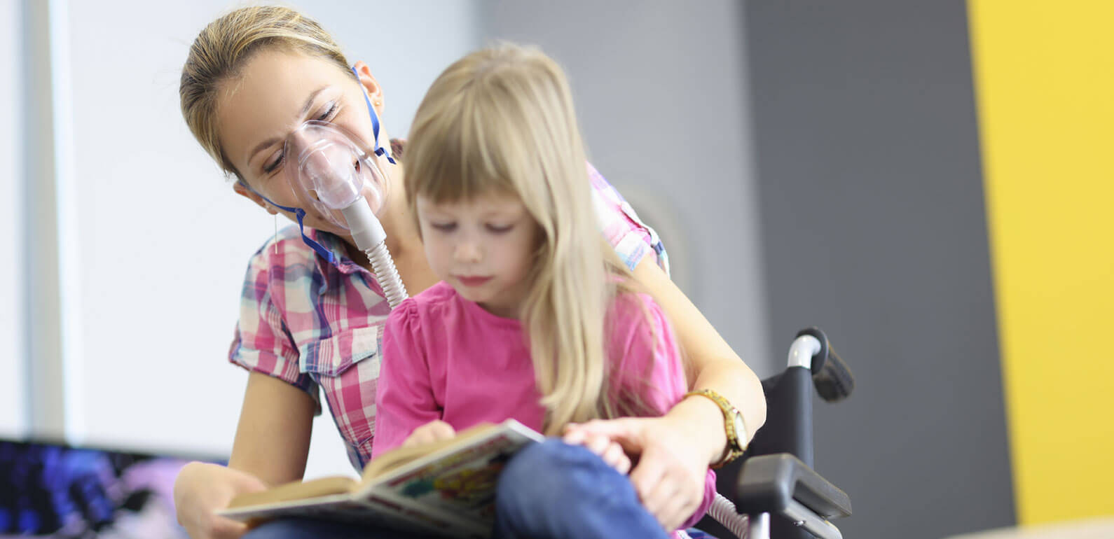 An adult female with oxygen support in a wheelchair with a child sat on her lap while reading