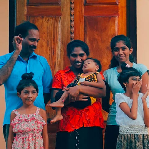 Sri Lankan family stood outside a family. A mother holding a baby next to her husband and three children