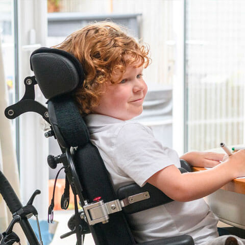 A young boy smiling in a wheelchair at a table writing